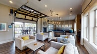This well-appointed billiards room in Gonzales, Louisiana, offers a thoughtfully designed social space that seamlessly blends indoor comfort with outdoor accessibility. The interior is anchored by rich wood-look flooring, crown molding, and a neutral color palette accented with bold yellow throw pillows that add a vibrant touch throughout.

The main lounge area features multiple groupings of plush, light-gray upholstered seating arranged around low-profile coffee tables, creating inviting conversation spaces. A central pool table serves as the room's focal point, illuminated by a striking cluster pendant light fixture overhead. Adjacent to the billiards area, a built-in bar ledge with barstool seating is complemented by wall-mounted flat-screen televisions, ideal for entertaining.

A floor-to-ceiling glass retractable garage door opens the space directly to a covered outdoor patio, flooding the interior with natural light and providing views of the surrounding community and pool area. The fireplace, clad in shiplap detailing, adds warmth and architectural character. A cozy lounge nook features textured wallcovering, built-in banquette seating, marble-top tables, and large windows dressed with sheer white curtains.

The covered outdoor patio extends the entertainment space with high-top tables and chairs overlooking the pool and lush landscaping, completing this versatile and dynamic amenity space.