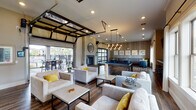 This well-appointed billiards room in Gonzales, Louisiana, offers a thoughtfully designed social space that seamlessly blends indoor comfort with outdoor accessibility. The interior is anchored by rich wood-look flooring, crown molding, and a neutral color palette accented with bold yellow throw pillows that add a vibrant touch throughout.

The main lounge area features multiple groupings of plush, light-gray upholstered seating arranged around low-profile coffee tables, creating inviting conversation spaces. A central pool table serves as the room's focal point, illuminated by a striking cluster pendant light fixture overhead. Adjacent to the billiards area, a built-in bar ledge with barstool seating is complemented by wall-mounted flat-screen televisions, ideal for entertaining.

A floor-to-ceiling glass retractable garage door opens the space directly to a covered outdoor patio, flooding the interior with natural light and providing views of the surrounding community and pool area. The fireplace, clad in shiplap detailing, adds warmth and architectural character. A cozy lounge nook features textured wallcovering, built-in banquette seating, marble-top tables, and large windows dressed with sheer white curtains.

The covered outdoor patio extends the entertainment space with high-top tables and chairs overlooking the pool and lush landscaping, completing this versatile and dynamic amenity space.
