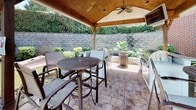 Located at 1100 Pulaski St in Columbia, South Carolina, this outdoor amenity space offers a well-appointed and thoughtfully designed environment for relaxation and social gatherings. The centerpiece of the space is a wood-framed covered pavilion, finished with a vaulted tongue-and-groove ceiling and equipped with a ceiling fan, providing a comfortable shaded retreat. Beneath the pavilion sits a fully outfitted outdoor kitchen, featuring granite countertops, a stainless steel grill, and ample prep space — ideal for al fresco dining and entertaining. High-top bar tables and mesh sling chairs are arranged throughout the covered and open patio areas, all set atop elegant stone pavers. Beyond the pavilion, the patio opens to a spacious courtyard anchored by a fire pit and surrounded by vibrant turquoise seating, leading to a resort-style swimming pool enclosed by wrought iron fencing. Tall palm trees, manicured hedgerows, decorative rock borders, and lush tropical plantings frame the entire outdoor area, enhancing its resort-like ambiance. The surrounding brick building architecture and stone retaining walls add a sense of permanence and sophistication to the overall setting, making this a standout outdoor living destination.