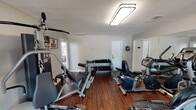 Located at 5450 Texas Ave in Abilene, Texas, this single-floor fitness center offers a comprehensive workout environment designed to accommodate a variety of exercise routines. The space is finished with rich, dark hardwood-style flooring that runs throughout, complementing the neutral gray walls and textured ceiling overhead. Bright flush-mount lighting ensures the room is well-illuminated at all times.

The fitness center is thoughtfully organized to maximize usability. A full wall of floor-to-ceiling mirrors lines one side of the room, visually expanding the space and allowing users to monitor their form during workouts. Cardio equipment is prominently represented, with treadmills, a recumbent bike, and an elliptical trainer from the True fitness brand positioned throughout the floor. A multi-use Paramount cable machine provides options for a wide range of strength-training exercises, while a tiered dumbbell rack stocked with free weights supports additional resistance training.

Large windows along one wall allow natural light to filter in, offering views of the surrounding outdoor greenery. A connected doorway leads to an adjacent space, and a designated employees-only door maintains operational boundaries. The overall ambiance is clean, functional, and inviting, making it a practical fitness resource for the community it serves.