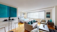 This thoughtfully designed studio is laid out across a single floor, making efficient use of its open-plan space. The main living area seamlessly integrates the sleeping and lounging zones, anchored by warm parquet wood flooring that runs throughout. A large bank of windows along one wall fills the room with abundant natural light, offering leafy outdoor views. The living area is generously sized to accommodate a full sofa seating arrangement alongside a bed, demonstrating excellent spatial flexibility. A compact bar-height table tucked against a partial wall creates a convenient dining nook.

A separate galley-style kitchen features white cabinetry, dark countertops, a stainless steel sink, and a full-size refrigerator. The black-and-white tile flooring adds a classic, crisp contrast to the bright white finishes. A small window provides additional ventilation and light to the space.

A dedicated walk-in closet with built-in shelving and a dresser offers ample storage. The full bathroom is finished with white subway tile walls, a combined tub and shower, a vanity with under-sink storage, and a large mirror. Throughout the unit, a cohesive teal and neutral color palette ties the spaces together, creating a calm and inviting ambiance.