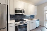 Located at 1100 Dahlia St NW in Washington, DC, this well-appointed one-bedroom, one-bathroom unit offers a thoughtfully designed open-concept layout. The combined living, dining, and kitchen area maximizes the efficient use of space, creating a bright and functional environment anchored by light hardwood-style flooring throughout. The kitchen is a standout feature, showcasing crisp white shaker cabinetry extending to the ceiling, a striking dark blue subway tile backsplash, quartz countertops, and a full suite of stainless steel appliances including a range, over-the-range microwave, refrigerator, and dishwasher. Track lighting overhead enhances the modern aesthetic, while a large window at the end of the living area invites natural light and outdoor views. The bedroom is a serene retreat, comfortably sized with its own window providing additional natural light. The bathroom is clean and contemporary, featuring a white shaker-style vanity with quartz countertop, an arched mirror, and a soaking tub with tile surround. A dedicated closet and in-unit laundry add practical convenience, rounding out this efficiently designed urban residence.