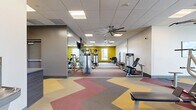 Located at 2157 E Apache Blvd in Tempe, Arizona, this single-floor fitness center spans 2,060 square feet and offers a well-equipped, thoughtfully designed exercise space. The facility is organized to accommodate both cardiovascular training and strength conditioning within an open layout.

The flooring throughout the space is a distinctive chevron-patterned design in bold shades of red, yellow, gray, and purple, adding visual energy to the environment. Accent walls painted in mustard yellow contrast with the neutral white walls, contributing to a bright and motivating atmosphere.

The cardio zone features multiple treadmills, elliptical machines, stair climbers, and stationary bikes positioned near floor-to-ceiling windows that allow ample natural light and outdoor views. Large ceiling fans are mounted overhead, promoting air circulation throughout the space.

The strength training area includes a variety of cable machines, weight benches, free weights, and multi-functional resistance equipment. Full-length mirrors line portions of the walls, enhancing the sense of space and allowing users to monitor their form.

Additional amenities include a water fountain station, a flat-screen television mounted on the wall, and a glass-enclosed corridor connecting to adjacent areas. Recessed and flush-panel ceiling lighting ensures consistent illumination throughout the facility.