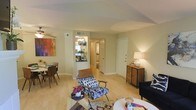 This model apartment in Richardson, Texas offers a well-planned single-floor layout designed for comfortable everyday living. The open-concept living and dining area serves as the heart of the home, featuring light hardwood-style flooring that extends throughout, creating a bright and cohesive atmosphere. Neutral wall tones provide a versatile backdrop, while warm ambient lighting from a ceiling fan fixture and table lamps adds a welcoming glow to the space. The living area is generously proportioned, offering ample room for furniture arrangement and entertaining. A decorative accent chair and layered area rug contribute to the stylish, mid-century modern aesthetic seen throughout the unit. Adjacent to the living space, the dining area accommodates a full dining set beneath a contemporary chandelier, perfect for everyday meals or hosting guests. A pass-through view into the kitchen reveals dark cabinetry and updated appliances, hinting at a functional and efficient cooking space. A hallway leads further into the unit, providing access to additional rooms and closet storage. The entryway is accented by a classic white door and a storage dresser, offering practical organization at the threshold. Overall, this model apartment presents a clean, stylish, and move-in-ready living environment in the heart of Richardson, TX.