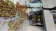 The entrance to Dwell at 2440 makes a distinct architectural statement, combining modern design elements with natural touches to create a welcoming and visually engaging arrival experience. The façade is clad in sleek dark tile, providing a refined backdrop for the prominent "2440" signage mounted above the entryway. A bold, angular glass canopy extends outward over the entrance, offering shelter while allowing natural light to filter through, lending the threshold a bright and open feel. Automatic sliding glass doors framed in dark aluminum provide a clean, contemporary transition point into the building's interior. One of the most striking features of the exterior is the living trellis structure positioned to the left of the entrance, where climbing vines cascade dramatically with rich seasonal foliage in deep reds, greens, and ambers. This organic element softens the hard architectural lines and introduces a natural warmth to the otherwise modern aesthetic. The surrounding pavement is composed of large-format tiles that reinforce the polished character of the building. Through the glass doors, the interior lobby is visible, hinting at a well-lit, refined space beyond. The overall ambiance of the entrance is one of contemporary urban sophistication balanced with thoughtful, nature-inspired design details.