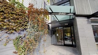 The entrance to Dwell at 2440 makes a distinct architectural statement, combining modern design elements with natural touches to create a welcoming and visually engaging arrival experience. The façade is clad in sleek dark tile, providing a refined backdrop for the prominent "2440" signage mounted above the entryway. A bold, angular glass canopy extends outward over the entrance, offering shelter while allowing natural light to filter through, lending the threshold a bright and open feel. Automatic sliding glass doors framed in dark aluminum provide a clean, contemporary transition point into the building's interior. One of the most striking features of the exterior is the living trellis structure positioned to the left of the entrance, where climbing vines cascade dramatically with rich seasonal foliage in deep reds, greens, and ambers. This organic element softens the hard architectural lines and introduces a natural warmth to the otherwise modern aesthetic. The surrounding pavement is composed of large-format tiles that reinforce the polished character of the building. Through the glass doors, the interior lobby is visible, hinting at a well-lit, refined space beyond. The overall ambiance of the entrance is one of contemporary urban sophistication balanced with thoughtful, nature-inspired design details.