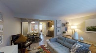 This single-floor residence offers a thoughtfully designed open-concept layout that seamlessly connects the living, dining, and kitchen areas into one cohesive and inviting space. The living area serves as the centerpiece, featuring a contemporary light-blue tufted sofa, a richly patterned area rug, and a warm floor lamp that contributes to the space's relaxed yet stylish ambiance. Decorative wall art and floating shelves add personality and a curated feel to the room. Flowing naturally from the living space, the dining area accommodates a round table with seating for multiple guests, anchored by a pendant light overhead — ideal for both casual meals and social gatherings. The kitchen is visible beyond the dining area and features dark wood cabinetry with light countertops, offering functional workspace in a compact yet efficient footprint. Wood-style flooring runs throughout, lending warmth and visual continuity across all areas. Neutral wall tones and consistent lighting create a bright, cohesive atmosphere throughout the home. The layout maximizes the use of space while maintaining a comfortable flow between zones, making it practical for everyday living.