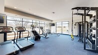 Located at 1415 County Road B W in Roseville, Minnesota, this fitness center is a well-appointed, single-floor facility designed to support a wide range of workout routines. The main exercise room is characterized by floor-to-ceiling windows along an entire wall, flooding the space with natural light and providing open views of the surrounding outdoor landscape — creating an energizing atmosphere for workouts.

The cardio zone is equipped with treadmills, a stair climber, and a stationary cycling bike, all positioned to take advantage of the expansive window views. A dumbbell rack with a full range of free weights and a weight bench provide ample options for strength training. A TRX suspension training system and a wall-mounted functional training rig stocked with kettlebells, medicine balls, and battle ropes add versatility for dynamic functional fitness routines.

An adjoining studio area features light hardwood flooring, large wall-to-wall mirrors, and racks holding a variety of stability balls and medicine balls — ideal for stretching, core training, or group exercise. Recessed lighting throughout ensures consistent illumination across both areas. Blue carpet tile flooring in the main exercise room adds a clean, modern finish. The overall design is contemporary and purposeful, offering a comprehensive fitness environment within a compact, efficient footprint.