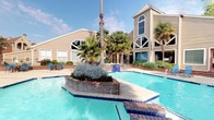 This outdoor pool area offers a well-appointed recreational retreat designed for relaxation and leisure. The centerpiece is a generously sized swimming pool with clear turquoise water, complemented by a separate hot tub/spa positioned adjacent to the main pool. A unique raised brick planter island adds a distinctive architectural touch to the pool's layout, adorned with tropical plants and palms that enhance the resort-like atmosphere.

The expansive concrete sun deck surrounds the pool and is furnished with multiple blue lounge chairs and additional seating, providing ample space for sunbathing and outdoor relaxation. Mature palm trees and lush greenery frame the perimeter, offering natural shade and a tropical ambiance. A dedicated patio dining area with tables and chairs is also visible, ideal for outdoor gatherings.

Access to the pool area is provided through a gated entry with wooden framed doors, reinforcing the secured and private nature of the space. Large glass sliding doors from an adjacent interior space overlook the pool, creating a seamless indoor-outdoor connection. The area is enclosed by a black iron fence bordered by mature trees, ensuring both privacy and an open, airy feel under the open sky.