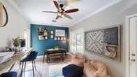 Located at 3214 Brassfield Rd in Greensboro, NC, this thoughtfully designed living room blends work, leisure, and social interaction into one cohesive space. The room is anchored by a striking teal accent wall adorned with framed artwork and rustic floating shelves, creating a bold focal point that adds depth and character. Crown molding runs along the perimeter of the ceiling, lending an elegant, finished look throughout.

A standout feature is the live-edge wood plank desk, which spans one full wall and is supported by angled wooden legs. Paired with navy blue upholstered bar stools and dual desktop monitors, this built-in workstation offers a functional yet stylish workspace. A wall-mounted flat-screen TV and an industrial-style media console anchor the opposite end of the room, complementing the warm hardwood-style flooring beneath.

The lounge area features plush bean bag chairs, a knit pouf ottoman, and a colorful area rug, creating a relaxed, inviting atmosphere. A large window fitted with horizontal blinds fills the space with natural light and provides views of the surrounding greenery. An oversized Scrabble board mounted on the wall adds a playful, social touch. Overhead, a five-blade ceiling fan with a cage-style light fixture ties the room's modern-rustic aesthetic together seamlessly.