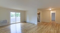 This single-floor residence offers a clean, functional layout with a bright and open living area as its centerpiece. The space is finished with light-toned hardwood flooring throughout, lending warmth and continuity across the rooms. Large sliding glass doors in the main living area flood the interior with natural light while providing direct access to an outdoor area, where lush greenery is visible beyond a privacy fence — a pleasant, verdant backdrop. A baseboard heating system runs along the wall, ensuring year-round comfort. The crisp white walls and neutral palette create a clean, versatile canvas well-suited to a variety of furnishing styles. Accessible directly from the living area, the kitchen is compact yet functional, featuring wooden cabinetry and a stainless steel refrigerator that adds a modern touch. The open sightlines between the living space and kitchen promote a connected, airy feel throughout. The overall ambiance of the space is light, understated, and move-in ready, offering a practical and comfortable environment with thoughtful details that balance simplicity with functionality.