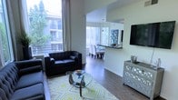 This three-floor space makes a strong impression with its open-concept design and generous sense of volume. The living area benefits from floor-to-ceiling windows that flood the room with natural light and offer views of lush greenery outside, creating a bright, airy atmosphere. Soaring ceilings add a dramatic vertical dimension to the space, enhancing its overall sense of openness.

The living room is anchored by a tufted dark leather sofa and loveseat arrangement, complemented by a sculptural glass-top coffee table resting on a yellow and grey geometric area rug. Rich hardwood flooring runs throughout, lending warmth and cohesion to the space. A wall-mounted flat-screen television and an ornate mirrored dresser add functional and decorative elements to the room.

The layout transitions smoothly into a dining area, where a rectangular table with upholstered white chairs is bathed in natural light from nearby windows. Adjacent to the dining space is a kitchen counter bar, topped in dark stone, with pendant lighting overhead and a decorative tile backsplash visible in the background. The overall ambiance is contemporary, polished, and welcoming.