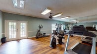 This fitness center is housed in a single-floor space designed to support a well-rounded workout experience. The room features warm hardwood flooring throughout, complemented by soft sage-green walls accented with white chair rail molding and crown molding — lending the space a polished, welcoming aesthetic uncommon in typical gym environments.

Natural light filters in through multiple windows and a set of classic white French doors, which serve as the main entrance and provide an open, airy feel. Flush-mounted fluorescent light fixtures and a ceiling fan ensure the space remains well-lit and comfortable during use.

A large floor-to-ceiling mirror lines one wall, visually expanding the room and allowing users to monitor their form during exercise. The equipment selection includes multiple treadmills, an elliptical trainer, a stationary recumbent bike, and a multi-function cable strength training machine, offering options for both cardiovascular and resistance training.

A built-in shelving unit with a wall-mounted television provides an entertainment feature for users during workouts. A posted fitness center rules board keeps the space organized and user-friendly. The textured ceiling adds a subtle architectural detail consistent throughout the room. Overall, the fitness center balances functionality with a refined interior design, creating an inviting and practical environment for exercise.