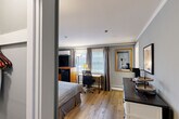 Located at 473 Main St in Barnstable, MA, this well-appointed one-bedroom, one-bathroom suite offers a thoughtfully organized layout across a single floor. The main room is finished with warm-toned hardwood flooring and soft gray walls accented by crisp crown molding, creating a clean and inviting atmosphere. Multiple windows dressed with draped curtains and horizontal blinds allow natural light to fill the space while providing privacy. The bedroom area comfortably accommodates a queen bed and includes a wooden media console with a flat-screen TV, a wall-mounted mini-split unit for climate control, and a compact mini-fridge and microwave for in-room convenience. A dedicated workspace with a desk and ergonomic chair is positioned near the windows, making the room functional for both leisure and productivity. A charming white barnwood-style console serves as a kitchenette station, complete with a dark countertop and open shelving, adding a touch of farmhouse character. A framed mirror with a gold accent and coastal-themed wall art contribute to the room's warm, curated aesthetic. The private bathroom features a clean, all-white design with a vanity, framed mirror, and bright overhead lighting, conveying a fresh and tidy feel throughout.
