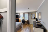 Located at 473 Main St in Barnstable, MA, this well-appointed one-bedroom, one-bathroom suite offers a thoughtfully organized layout across a single floor. The main room is finished with warm-toned hardwood flooring and soft gray walls accented by crisp crown molding, creating a clean and inviting atmosphere. Multiple windows dressed with draped curtains and horizontal blinds allow natural light to fill the space while providing privacy. The bedroom area comfortably accommodates a queen bed and includes a wooden media console with a flat-screen TV, a wall-mounted mini-split unit for climate control, and a compact mini-fridge and microwave for in-room convenience. A dedicated workspace with a desk and ergonomic chair is positioned near the windows, making the room functional for both leisure and productivity. A charming white barnwood-style console serves as a kitchenette station, complete with a dark countertop and open shelving, adding a touch of farmhouse character. A framed mirror with a gold accent and coastal-themed wall art contribute to the room's warm, curated aesthetic. The private bathroom features a clean, all-white design with a vanity, framed mirror, and bright overhead lighting, conveying a fresh and tidy feel throughout.