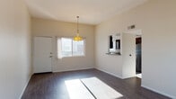 This single-floor residence offers a straightforward and functional layout designed for comfortable everyday living. The main living area is open and generously proportioned, finished with rich, dark hardwood-style flooring that runs throughout the space. Neutral beige walls and vaulted ceilings contribute to a bright and airy atmosphere, while a stylish gold pendant light serves as a focal point, adding a warm decorative accent to the room. Natural light streams in through a front-facing window, casting a natural glow across the floors and enhancing the overall sense of openness.

A pass-through window connects the living area to the kitchen, providing a convenient and sociable link between the two spaces. The kitchen features dark cabinetry and is equipped with a stainless steel refrigerator, visible through the adjoining doorway. The layout, as seen in the dollhouse view, also includes a dedicated bedroom and a bathroom. The bedroom continues the dark hardwood-style flooring and benefits from natural light. The bathroom is clean and functional, featuring a full-size bathtub with a handheld showerhead and white tile surround. Overall, the space presents a clean, well-maintained interior with a cohesive neutral palette.