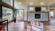 This single-floor space makes a strong architectural impression through its dramatic double-height ceilings and expansive floor plan. Rich hardwood flooring runs throughout, anchoring the open layout and complementing the warm, neutral wall tones that define the overall aesthetic.

The main lounge area serves as the focal point of the space, centered around a striking stacked-stone fireplace with a rustic wood mantel. A wall-mounted television sits above the mantel, while comfortable upholstered seating arranged around a patterned area rug creates a welcoming gathering area. Decorative wall sconces provide warm ambient lighting throughout.

Large, multi-pane windows flood the space with natural light and offer views of the surrounding greenery. A bar-height communal table accompanied by vibrant orange metal stools is positioned near the windows, providing an informal seating area that takes full advantage of the outdoor views and abundant daylight.

An adjacent room, visible through a wide doorway, extends the usable area and features additional seating, storage furnishings, and wall art. The overall ambiance is modern yet warm, blending natural materials with contemporary design elements for an inviting, well-appointed environment.