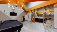 Located at 3470 Ft Independence St in the Kingsbridge neighborhood of the Bronx, New York, this single-floor community amenity space delivers a bold, energetic atmosphere defined by its striking orange-accented ceiling beams, high ceilings, and light gray tile flooring throughout. The open-plan game and recreation area serves as the centerpiece, featuring a full-size pool table, a classic Pac-Man arcade cabinet, and a mid-century modern sputnik chandelier that adds a retro flair. Vibrant wall art and eclectic decor, including music-themed posters and sculptural wall pieces, give the space a distinctive personality. A decorative gold lattice room divider elegantly separates zones while maintaining visual openness. The adjacent lounge area offers a large black leather sectional sofa, a colorful area rug, and a fireplace topped with a large flat-screen TV, creating a warm and inviting gathering spot. A cozy private meeting nook features a solid wood conference table surrounded by teal upholstered chairs, pendant lighting, and exposed red piping overhead, ideal for small group gatherings. The space also includes a kitchenette area with a refrigerator, completing this well-rounded, thoughtfully designed community hub.