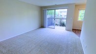This single-story space features a generously sized living area finished with soft, wall-to-wall carpeting that adds warmth and comfort underfoot. The room is bathed in natural light, courtesy of a large sliding glass door that serves as both a functional entry point and a visual focal point. The door opens onto a private balcony enclosed by a classic railed barrier, offering views of lush greenery and surrounding landscaping. The neutral, light beige walls provide a clean and versatile backdrop, complementing the bright and airy atmosphere throughout. Vertical blinds frame the sliding door, offering flexible light control and privacy. The smooth ceiling with subtle texture and clean white baseboards complete the well-maintained interior. The open layout of the living space provides ample room for a variety of furniture arrangements, making it highly adaptable to different lifestyle needs. Overall, the space strikes a balance between simplicity and functionality, with its connection to the outdoors and abundance of natural light serving as defining characteristics of the interior's overall ambiance.