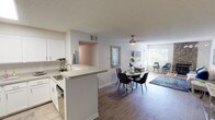 This single-floor residence offers a thoughtfully designed open-concept layout that connects the kitchen, dining, and living areas into one cohesive, functional space. The kitchen is outfitted with white shaker cabinetry, light quartz countertops, stainless steel appliances — including a refrigerator, range, and microwave — and a peninsula that provides additional prep space and a natural division from the dining area. The dining space sits beneath a ceiling fan and is illuminated by natural light streaming through large windows, creating a bright and welcoming atmosphere throughout. The living room is a standout area, anchored by a striking floor-to-ceiling stone fireplace that adds warmth and architectural character. Wood-look flooring runs through the main living areas, while the generously sized bedroom features plush carpet and ample natural light. The bathroom is clean and modern, featuring white cabinetry, a wide vanity with a large mirror, and updated fixtures. Throughout the residence, a neutral color palette of whites and soft grays provides a versatile backdrop, while textured ceilings add a classic touch. The layout, as seen in the dollhouse view, maximizes the use of space with a well-organized bedroom that includes a walk-in closet area.