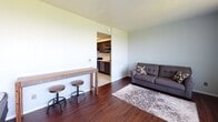 Located at 100 Fairview Square in Ithaca, NY, this single-floor unit offers a practical and comfortable layout designed for efficient everyday living. The open-concept living area features rich dark hardwood flooring that runs throughout, neutral light-toned walls, and a large picture window fitted with vertical blinds that floods the space with natural light and frames a pleasant view of the surrounding greenery. The living area provides ample room for seating and casual dining, with the layout flowing naturally into the adjoining kitchen. The kitchen is well-equipped with dark cabinetry, marble-style countertops, stainless steel appliances including a gas range, built-in microwave, and dishwasher, complemented by light vinyl flooring that contrasts cleanly with the darker finishes. A dedicated entry door area is conveniently accessible from the kitchen. The bedroom is a quiet, carpeted retreat with dark hardwood flooring, clean white walls, and enough space to comfortably accommodate a full-sized bed and additional furnishings. Throughout the unit, the design emphasizes clean lines, functional storage, and a cohesive neutral palette, making it a versatile and move-in ready space in the heart of Ithaca.