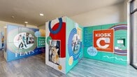 This 700 square foot amenity space at The Flats at Palisades showcases a blend of functionality and artistic expression. The area is characterized by its vibrant, mural-covered walls that display colorful graphics, motivational phrases, and branding elements. These murals create a lively and energetic atmosphere throughout the space.

The flooring consists of durable, wood-like planks that provide a warm contrast to the white walls and ceiling. Recessed lighting fixtures are evenly spaced across the ceiling, ensuring ample illumination throughout the area.

A notable feature is the package locker area, indicated by a series of uniform doors along one wall. This practical amenity allows residents to securely receive deliveries at their convenience. Large windows near the entrance allow natural light to flood the space, creating an open and airy feel.

The layout includes several freestanding structures adorned with murals, which may serve as information kiosks or additional package lockers. These structures add depth and visual interest to the space while potentially offering additional functionality.

The overall design of this amenity area strikes a balance between practicality and aesthetic appeal, creating a welcoming environment for residents that reflects a modern, community-oriented living experience.