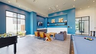This contemporary common living space makes a striking first impression with its bold cobalt blue accent walls contrasted against crisp white wainscoting and neutral-toned large-format tile flooring. The room is anchored by a comfortable gray sectional sofa, a rustic wooden coffee table, and a pair of leather accent chairs, all arranged atop a vibrant abstract area rug that adds a dynamic layer of color and texture. Mounted wall-mounted shelving in warm wood tones displays decorative objects and artwork, while a flat-screen TV and a dark wood credenza complete the media-ready lounge area.

One of the space's most notable architectural features is its soaring ceiling height, which opens dramatically toward a bank of tall, black-framed windows that flood the room with natural light and offer views of the surrounding greenery. Recessed lighting and a modern chandelier provide warm, layered illumination throughout.

The space is further equipped for recreation, featuring a full-size pool table and a foosball table, making it well-suited for social gatherings and leisure. The entry area is defined by sleek black steel-framed glass doors that lend an industrial-modern aesthetic. Overall, the space strikes a confident balance between lively design and functional versatility.