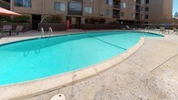 This outdoor recreational area serves as a central amenity for a multi-story residential complex, offering a well-appointed pool and surrounding deck space designed for both relaxation and socializing. The centerpiece is a generously sized swimming pool with clear turquoise water, curved edges, and metal entry rails, set within a wide concrete surround that provides ample room for sunbathing and leisure. The sundeck is furnished with slatted lounge chairs and a wooden bench table, ideal for unwinding in the open air. A designated dining area features a round glass-top table paired with sling chairs, shaded by a large orange market umbrella, creating a comfortable outdoor gathering spot. The pool area is enclosed by a combination of metal railing fencing and a wooden gate, with posted safety and health signage prominently displayed at the entrance. Lush shrubs and greenery line the perimeter, softening the texture-coated exterior walls of the surrounding multi-story buildings. String lights are strung above the deck, suggesting an inviting atmosphere during evening hours. A vending machine is conveniently positioned near the pool entrance. The space is entirely at ground level, offering easy accessibility and an open, sun-filled ambiance throughout the day.