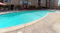 This outdoor recreational area serves as a central amenity for a multi-story residential complex, offering a well-appointed pool and surrounding deck space designed for both relaxation and socializing. The centerpiece is a generously sized swimming pool with clear turquoise water, curved edges, and metal entry rails, set within a wide concrete surround that provides ample room for sunbathing and leisure. The sundeck is furnished with slatted lounge chairs and a wooden bench table, ideal for unwinding in the open air. A designated dining area features a round glass-top table paired with sling chairs, shaded by a large orange market umbrella, creating a comfortable outdoor gathering spot. The pool area is enclosed by a combination of metal railing fencing and a wooden gate, with posted safety and health signage prominently displayed at the entrance. Lush shrubs and greenery line the perimeter, softening the texture-coated exterior walls of the surrounding multi-story buildings. String lights are strung above the deck, suggesting an inviting atmosphere during evening hours. A vending machine is conveniently positioned near the pool entrance. The space is entirely at ground level, offering easy accessibility and an open, sun-filled ambiance throughout the day.