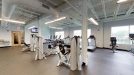 This expansive fitness center is thoughtfully organized into distinct workout zones, offering a comprehensive range of equipment to support diverse training needs. The facility is characterized by its open, industrial-influenced design, featuring an exposed ceiling structure with white-painted steel beams, ductwork, and track lighting that collectively contribute to a bright, functional atmosphere.

The cardio zone lines one wall beneath a row of large windows, housing multiple treadmills, upright stationary bikes, and elliptical machines. Natural light filters generously through these windows, creating an energizing environment for aerobic workouts. A wall-mounted television provides additional entertainment options in this area.

The strength training section occupies the central and adjacent floor space, featuring an array of selectorized cable machines and weight stations arranged to allow comfortable circulation between equipment. A dedicated free-weight area includes a Smith machine with weight plates, flat benches, and additional free-weight accessories. Full-length mirrors along select walls enhance spatial awareness during workouts.

Throughout the facility, durable speckled rubber flooring provides both safety and resilience underfoot. Structural columns are integrated into the layout without impeding traffic flow. The light gray walls and abundant overhead lighting maintain a clean, professional aesthetic, making this fitness center a well-rounded and functional training environment.