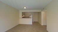 This single-floor residence presents an open and functional layout, with the living area and kitchen connected through a half-wall pass-through that adds visual separation while maintaining an open feel. The living area is finished with wall-to-wall carpeting in a neutral beige tone, contributing to a warm and understated ambiance throughout the space.

The kitchen is positioned toward the rear and features light-toned cabinetry lining the walls, providing ample storage. A white refrigerator is visible, along with standard countertop surfaces. The pass-through counter serves as a casual divider between the kitchen and the main living space, adding practicality for everyday use.

The ceilings are finished with a textured surface and fitted with a central flush-mount light fixture that illuminates the room. Neutral beige walls are consistent throughout, creating a cohesive and clean aesthetic. A visible HVAC vent is integrated into the ceiling, and doorways lead to additional rooms, suggesting a straightforward and efficient floor plan. The overall ambiance is calm and move-in ready, suited for functional everyday living.