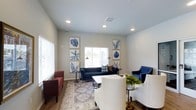 This single-floor space offers a thoughtfully designed interior that balances comfort and style. The living area serves as the heart of the space, featuring recessed ceiling lighting that casts a warm, even glow throughout the room. Large windows allow abundant natural light to fill the area, enhancing the open and airy atmosphere created by the elevated ceilings and clean architectural lines.

The walls are finished in a soft, neutral tone that provides a versatile backdrop for the carefully curated furnishings. Seating arrangements include a deep navy blue sofa paired with a coordinating accent chair, a rich burgundy armchair, and cream-upholstered seating that together create a welcoming, layered aesthetic. A white coffee table anchors the central seating area atop a classic patterned area rug laid over hardwood flooring.

Decorative botanical wall art in blue and white tones is symmetrically displayed on either side of the main window, adding a refined visual element to the space. Warm-toned floor lamps complement the recessed lighting and contribute to the inviting ambiance. Overall, this space presents a clean, well-maintained interior that is both functional and visually cohesive, offering a comfortable environment suited for relaxation or social gatherings.