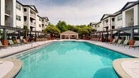 Located at 25821 Burbage Circle in Cary, NC, this beautifully designed outdoor amenity space serves as the centerpiece of a multi-story residential community. The courtyard is framed on multiple sides by three-story buildings featuring white siding, brick accents, and private balconies, creating an enclosed, resort-like atmosphere.

The focal point of the space is a generously sized swimming pool with a smooth, curvilinear design and crystal-clear blue water. Flanking the pool on both sides are extended sundecks lined with lounge chairs, offering ample room for relaxation. At the far end of the pool, a covered cabana pavilion provides a shaded gathering area, complete with a television and seating — ideal for outdoor entertaining.

Surrounding the pool deck are well-maintained raised garden beds constructed with classic red brick walls, filled with a rich variety of lush shrubs, ornamental grasses, and flowering plants. Black iron fencing and tall columnar evergreen trees add vertical interest and a sense of privacy. Accessible ramps with metal railings ensure seamless connectivity throughout the courtyard.

Outdoor dining areas with umbrella-shaded tables and chairs are positioned along the perimeter, offering comfortable spaces for al fresco dining. The combination of manicured landscaping, open sky views, and cohesive brick-and-siding architecture creates a welcoming and vibrant community outdoor environment.