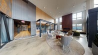 This thoughtfully designed single-floor amenity space makes a striking first impression with its grand, double-height lobby entrance, framed by expansive floor-to-ceiling windows that flood the interior with natural light and offer views of the surrounding streetscape. Polished concrete floors run throughout, anchoring the contemporary aesthetic.

The main lounge area centers around a dramatic floor-to-ceiling fireplace clad in rich wood paneling, complemented by a built-in log storage niche. Carefully curated seating arrangements — including sculptural lounge chairs, a plush green sofa, and leather accent pieces — are organized on layered area rugs, creating an inviting social atmosphere.

A café bar area features patterned tile flooring, warm wood cabinetry, pendant lighting, and a retro blue refrigerator as a playful focal point. Adjacent dining nooks offer tufted banquette seating alongside bold, colorful wall art and pendant-lit booth seating for a more intimate dining experience.

A secondary lounge alcove showcases a striking graphic mural accent wall, dark ceiling detail, and warm mid-century furnishings. A fully equipped community kitchen provides stainless steel appliances, a cooktop, oven, and open shelving. Private pods offer quiet, bookable spaces for focused work or meetings, completing this richly detailed, multi-functional communal environment.
