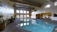 This indoor aquatic facility offers a welcoming and well-appointed environment centered around a generously sized swimming pool. The pool itself features underwater lighting that gives the water a clear, inviting blue-green hue, complemented by a textured, slip-resistant deck surrounding the perimeter. The space is architecturally distinguished by soaring ceilings with an exposed structural beam system, lending an industrial-chic aesthetic to the interior. A dramatic wall of large, multi-pane windows floods the space with abundant natural light, offering views of the lush greenery outside and creating a seamless connection between the indoor and outdoor environments. The upper portion of the room features additional clerestory windows, further enhancing the bright and airy ambiance. Along the poolside, lounge seating is available for relaxation, while potted tropical plants add a touch of warmth and natural character to the setting. A separate enclosed area, likely housing changing rooms or additional amenities, is visible toward the rear of the facility. Wall-mounted screens are also present, adding a modern, functional element to the space. Overall, the facility combines practical aquatic infrastructure with a refined, comfortable atmosphere.
