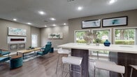 This single-floor communal space is designed with a contemporary aesthetic, combining functionality with modern style throughout its open layout. Warm-toned gray walls, wood-look plank flooring, and textured ceilings with recessed lighting create a cohesive and inviting atmosphere across all areas.

The lounge area serves as a central gathering space, featuring mid-century modern teal accent chairs, a tufted ottoman-style coffee table, and a patterned area rug anchoring the seating arrangement. An electric fireplace with a glowing red display is mounted to the wall, flanked by framed artwork and decorative panels, adding both warmth and visual interest. Multiple windows allow abundant natural light and provide views of the surrounding green landscape.

Adjacent to the lounge is a high-top communal table with a white surface and dark wood base, surrounded by sleek white bar-height stools. A textured mosaic tile accent wall provides a refined backdrop to this seating area, which also benefits from large windows overlooking lush outdoor greenery.

A dedicated computer workstation room offers a long built-in desk accommodating multiple desktop computers, providing a practical workspace within the facility. The room maintains the same neutral gray color palette and wood-look flooring found throughout the space.