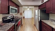 This single-floor residence offers a practical and comfortable layout designed for everyday living. The living area provides a welcoming atmosphere, furnished with leather seating and accented by warm, decorative touches including seasonal wreaths and framed wall art. A window unit provides climate control, while natural light filters in through blinds and curtains, adding a cozy warmth to the space. The entry area features light hardwood-style flooring that extends through the main living zones, creating a cohesive and clean aesthetic throughout.

The galley-style kitchen is a standout feature of the home, lined on both sides with rich, dark cherry wood cabinetry offering generous storage space above and below the counters. Granite-look countertops provide a durable and attractive work surface, complemented by a full suite of stainless steel appliances including a refrigerator, electric range, built-in microwave, and dishwasher. A double basin sink sits beneath a window, and a large double-door pantry or closet adds additional storage. A ceiling fan overhead and the continuation of hardwood-style flooring tie the kitchen seamlessly to the rest of the home, contributing to an overall atmosphere that is functional, tidy, and inviting.