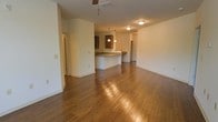 This single-story space is defined by its open-concept layout, seamlessly connecting the living area and kitchen into one cohesive, flowing environment. Rich hardwood flooring spans the entirety of the main area, reflecting light and adding warmth to the neutral, cream-toned walls throughout.

The kitchen is positioned at the back of the open floor plan and features a peninsula-style counter with dark granite or stone countertops, providing both workspace and a casual dining or seating option. Medium-toned wood cabinetry lines the kitchen walls, complementing the warm flooring below. A pair of pendant lights hangs above the peninsula, offering focused task lighting, while a flush-mount ceiling fixture provides additional ambient illumination to the kitchen zone.

The expansive living area benefits from a ceiling fan, promoting air circulation throughout the space. The open layout creates a generous sense of scale, making the area feel both airy and versatile. Doorways visible along the perimeter suggest additional rooms beyond the main living space. Overall, the space presents a practical and inviting interior with consistent, warm finishes throughout.