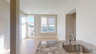 This single-floor space is designed with an open, flowing layout that connects the kitchen and living areas seamlessly. The kitchen serves as a centerpiece, anchored by a substantial granite island with an undermount sink and elegant veining throughout. Rich, medium-toned wood cabinetry extends to impressive heights beneath soaring ceilings with recessed lighting, while stainless steel appliances—including a French door refrigerator, electric range, and built-in microwave—complete the well-appointed culinary space. Light hardwood flooring runs throughout the main living areas, adding warmth and continuity.

The living area benefits from generous natural light through large windows fitted with roller shades, offering views of a neighboring urban streetscape. A glass-panel door provides access to an outdoor area, further enhancing the connection between interior and exterior. Bedroom spaces are calm and neutral, with the same light hardwood flooring and clean, minimal trim work.

The bathroom is particularly refined, featuring a floating vanity with granite countertops, white shaker-style cabinetry, and a large frameless mirror with flanking sconce lighting. Distinctive hexagonal floor tile and a tiled walk-in shower with glass enclosure add a polished, spa-like character. The overall ambiance throughout the space is clean, contemporary, and thoughtfully finished.