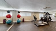 This well-equipped fitness center at 569 Little River Loop in Altamonte Springs, Florida, offers a comprehensive workout environment spread across a single floor. The space is designed to accommodate a wide range of exercise routines, with distinct areas dedicated to cardio, strength training, and functional fitness. Durable rubber flooring covers the entire facility, providing traction and impact absorption throughout. Large wall-mounted mirrors line multiple sections of the room, visually expanding the space and allowing users to monitor their form during workouts. Natural light filters in through several windows fitted with blinds, complemented by recessed ceiling lighting for consistent illumination at all hours. The cardio section is outfitted with a treadmill, elliptical trainer, and a stationary recumbent bike. A multi-station cable and weight machine anchors the strength training area, accompanied by a full rack of dumbbells in varying weights. Stability and exercise balls on a dedicated rack support flexibility and core training. Wall-mounted televisions provide entertainment, and posted fitness center rules ensure a structured environment. A glass-panel door offers direct exterior access, bringing in views of the surrounding greenery. The textured ceiling and neutral wall tones give the space a clean, practical aesthetic suited for daily fitness use.