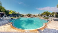 Situated at 20080 West Dixie Highway in Miami, Florida, this outdoor amenity space showcases a generously sized, resort-style swimming pool as its central feature. The pool features a sweeping curved design with crystal-clear turquoise water, and depth markers indicating a 4-foot depth, making it accessible for a range of swimmers. The pool deck is finished with warm-toned paver tiles that complement the South Florida aesthetic and provide ample space for lounging along the water's edge.

Surrounding the pool, lush tropical palm trees frame the area, reinforcing the resort-like ambiance characteristic of Miami living. Lounge chairs and seating areas are arranged along the perimeter, offering comfortable spots to relax in the abundant Florida sunshine. Shade umbrellas are positioned throughout the deck area, providing relief from the sun when needed. Elegant lamp posts line the space, ensuring the area is well-lit during evening hours.

In the backdrop, a multi-story residential building with a classic Mediterranean-inspired facade provides a striking visual anchor to the setting. The combination of the vibrant blue pool water, swaying palms, and clear blue skies creates a bright, welcoming outdoor retreat that captures the essence of Florida's warm, sun-soaked lifestyle.
