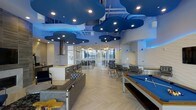 This dynamic amenity lounge is a single-floor space defined by its striking modern design and bold aesthetic. The most eye-catching architectural feature is the sculptural ceiling, adorned with oversized cloud-shaped acoustic panels in deep blue, complemented by recessed lighting and exposed industrial elements above. Blue accent lighting runs along the walls, reinforcing the space's vibrant, cohesive color palette.

The open floor plan is laid out across light-toned herringbone wood flooring, which adds warmth and texture to the otherwise contemporary environment. A billiards table with a blue felt surface anchors one side of the room, while a comfortable lounge area with upholstered seating and accent tables provides a relaxed gathering space nearby. A kitchenette with counter seating offers a convenient prep and serving area, ideal for entertaining.

Floor-to-ceiling glass partitions divide a secondary seating zone, furnishing it with industrial-style chairs and tables for a more structured setting. Natural light filters through the expansive glazing, brightening the interior and creating a sense of openness. Together, the recreational, social, and functional zones make this lounge a well-rounded, visually impressive space suited for community gatherings and leisure activities.