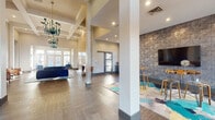 Located at 16401 Chenal Valley Dr in Little Rock, Arkansas, this single-floor community clubhouse exudes a refined and welcoming atmosphere. The space is defined by soaring ceilings adorned with coffered detailing and dramatic teal chandelier fixtures that serve as striking focal points throughout. Large floor-to-ceiling windows and glass entry doors flood the interiors with abundant natural light, highlighting the rich hardwood-style flooring that flows seamlessly across the space.

The main lounge area features a tufted navy velvet sofa, colorful accent chairs in teal and cognac leather, and vibrant area rugs that add layers of texture and color. A sleek linear fireplace set against a light wood-paneled accent wall anchors one end of the lounge, creating a cozy retreat. Opposite the lounge, a textured stone-inspired feature wall hosts a large flat-screen television alongside bar-height seating — ideal for social gatherings.

A glass-partitioned office or leasing area provides transparency while maintaining a professional, semi-private workspace. The clubhouse also includes a refreshment station with cabinetry and coffee service, positioned between dramatic metallic accent walls that lend an industrial-chic contrast to the space. Throughout, curated artwork and thoughtful furnishings contribute to a cohesive, upscale aesthetic.