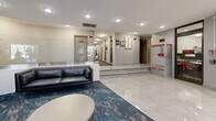 Located at 30 Godstone Road in Toronto's M2J neighbourhood, this residential lobby serves as an elegant and welcoming point of entry for the building. The space is finished with large-format marble-effect porcelain floor tiles that lend a clean, polished aesthetic throughout. A central lounge area anchors the lobby, featuring a sleek black leather sofa, a round coffee table, and a bold blue abstract area rug that adds a striking visual contrast to the light surroundings. The focal wall showcases a long linear electric fireplace with vivid colour-changing flames, flanked by framed landscape artwork and a classic wall clock, creating a warm and inviting atmosphere. Recessed pot lighting illuminates the space evenly, complementing the textured ceiling finish. A glass-railed raised platform defines a secondary zone within the lobby, housing a decorative plant that adds a natural touch. Toward the entrance, a Canada Post parcel locker and mailbox station provide convenient mail access for residents, positioned near the glazed entry doors. A decorative niche near the entrance displays a floral arrangement, adding a refined finishing detail. The lobby connects to building corridors via a short staircase, with clearly marked signage and fire safety equipment integrated throughout.