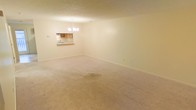 This multi-floor space spans three levels and presents a functional, straightforward layout. The main living area is finished with wall-to-wall carpet in a neutral beige tone, creating a warm, understated foundation throughout the room. Cream-colored walls contribute to a bright, open atmosphere, while a textured ceiling adds subtle visual detail overhead.

A chandelier serves as the central lighting fixture in the living area, providing ambient illumination and a modest decorative focal point. One of the notable architectural features of this space is a pass-through window connecting the living area to the kitchen, offering a sense of openness between the two zones without fully removing the wall separation. The kitchen, visible through the pass-through, features wooden cabinetry in a medium brown finish and includes a sink positioned beneath the window opening.

The overall ambiance of the space is calm and neutral, with a clean palette that offers flexibility for a variety of interior arrangements. The three-floor layout suggests additional living, sleeping, or utility spaces beyond what is visible, providing a meaningful sense of depth and versatility throughout the property.