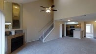 This two-story residence is defined by its striking vaulted ceilings in the main living area, creating an expansive, airy atmosphere that immediately draws the eye upward. A ceiling fan with a light fixture is centrally mounted, providing both airflow and ambient lighting to the generous living space below. Neutral-toned carpet covers the living area floor, contributing to a warm and understated aesthetic throughout.

A fireplace with a tiled surround anchors one wall of the living room, serving as a functional and visual focal point. A staircase with white-painted balusters and a light wood railing ascends from the main level to the upper floor, seamlessly integrated into the open layout.

Adjacent to the living area, the kitchen is visible through an open passage and features wood-toned cabinetry, black appliances including a range and overhead microwave, and overhead fluorescent lighting. The kitchen's practical layout and connection to the main living space make it well-suited for everyday use.

The two-level design offers a clear separation between the communal living spaces on the ground floor and the upper level, providing a functional and well-organized floor plan overall.