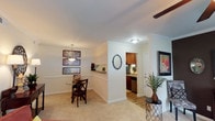This single-story residence offers a thoughtfully arranged open floor plan that seamlessly connects the primary living areas. Crown molding throughout adds a refined architectural detail, while neutral-toned walls create a bright and inviting atmosphere.

The dining area is highlighted by an elegant pendant chandelier that hangs above a glass-top dining table, complemented by framed wall art arranged in a gallery-style display. A half-wall partition subtly separates the dining space from the adjoining areas, adding structural interest without compromising the open feel.

The kitchen, accessible through a dedicated entryway, features warm wood cabinetry, dark appliances including a dishwasher, and hardwood-style flooring that contrasts with the plush carpeting found in the surrounding living spaces. A circular mirror adorns the wall near the kitchen entrance, adding a decorative focal point.

The living area is anchored by a ceiling fan, ensuring year-round comfort. A decorative console table with a potted plant and framed artwork further enhances the space's welcoming character. Overall, the layout balances functionality with comfortable, classic design.