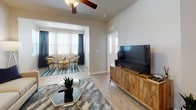 This single-story residence offers a well-designed, open layout that connects its primary living spaces seamlessly. The kitchen is a standout feature, outfitted with light wood-toned cabinetry, stainless steel appliances including a refrigerator, range, and microwave, and a generous white quartz peninsula with pendant lighting overhead. Light wood-style flooring runs throughout the kitchen and main living areas, contributing to a bright, cohesive aesthetic.

The living room provides a comfortable gathering space with light-filled windows fitted with blinds and drapes, a ceiling fan, and wood-style flooring. It opens to a dedicated sitting or sunroom area framed by large windows, allowing ample natural light. A separate dining nook accommodates a round table with seating for four, accented by warm neutral walls.

The bedroom features plush carpeting, multiple windows that invite natural light, a ceiling fan, and generous wall space. The bathroom is appointed with a large tub and shower combination, a vanity with stone-style countertops, built-in shelving, and tile flooring, offering a clean, functional design throughout. The overall ambiance of the space is warm, neutral, and move-in ready.