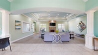 This single-floor community space offers a well-appointed, open layout designed for comfort and functionality. Upon entering, large format tile flooring and soft sage and teal-toned walls set a welcoming tone throughout. Elegant white decorative columns frame the entrance area, which features a glass-top table and seating, ideal for meetings or private conversations.

The main lounge area is generously sized, showcasing a striking floor-to-ceiling stone fireplace topped with a wall-mounted television as a central focal point. Plush grey sofas and accent chairs are arranged around a wood coffee table atop a vibrant blue-and-grey area rug, creating an inviting gathering space. Recessed lighting and a tiered tray ceiling with crown molding add architectural depth, while multiple windows flood the room with natural light.

A secondary lounge area features vaulted ceilings, additional seating, and tasteful wall art, extending the social atmosphere. The space transitions smoothly between tile and patterned carpet flooring throughout, delineating areas while maintaining a cohesive aesthetic. A small dining area with blue accent chairs adds a casual, functional touch. Overall, the space exudes a bright, airy ambiance with well-coordinated design elements that balance professionalism with relaxed comfort.