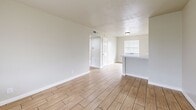 This 2-bedroom, 1-bathroom unit is designed with a practical and efficient single-floor layout. Wood-look tile flooring runs consistently throughout the space, lending warmth and visual continuity from room to room. The living room is bright and open, featuring a large window with blinds that invites natural light and offers outdoor views of surrounding greenery. A half-wall peninsula separates the living area from the kitchen, creating a semi-open feel. The kitchen is equipped with warm oak cabinetry, a double stainless steel sink with white countertops, and a gas range, providing a functional cooking space with its own window and direct access door. Both bedrooms are finished with the same wood-look tile flooring, neutral walls, and ample natural light from windows dressed with horizontal blinds. The second bedroom features low-profile built-in drawers along the baseboard, adding a practical storage element. The bathroom is a standout feature, showcasing a soaking tub and shower combination surrounded by marble-style wall panels and accented with a decorative band of mixed black, white, and iridescent mosaic tile. A window above the tub allows natural light into the space. A hallway connects the bedrooms and bathroom, completing the well-organized floor plan.